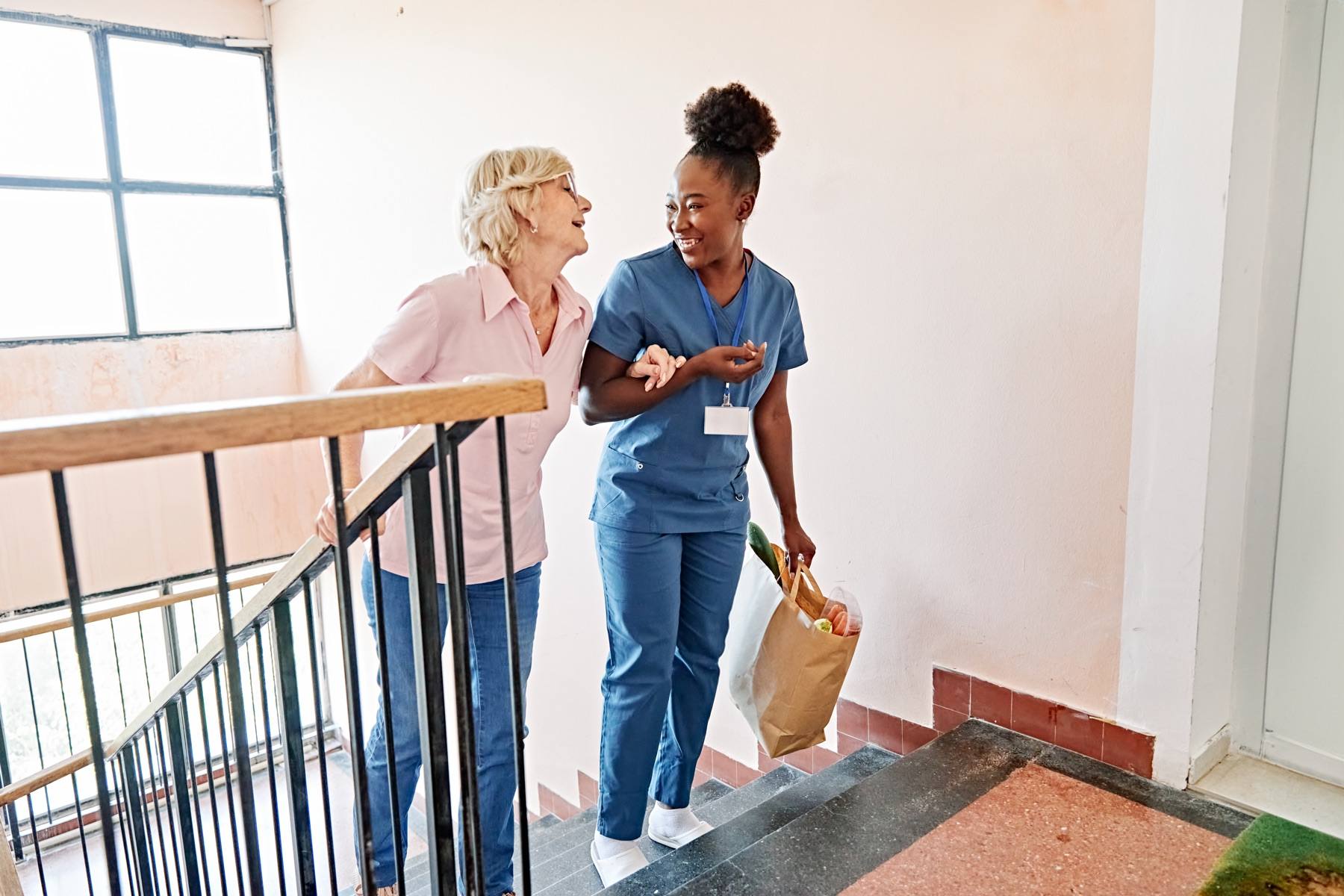 A caregiver walks up a flight of stairs arm in arm with an older woman, carrying her bag of groceries.