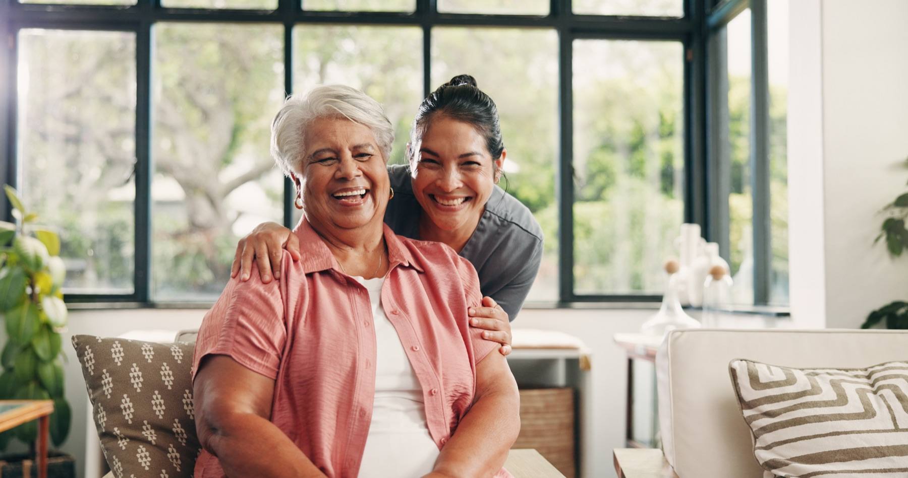 A smiling caregiver stands behind an older woman with her arms around her shoulders, both laughing, in a bright living room.