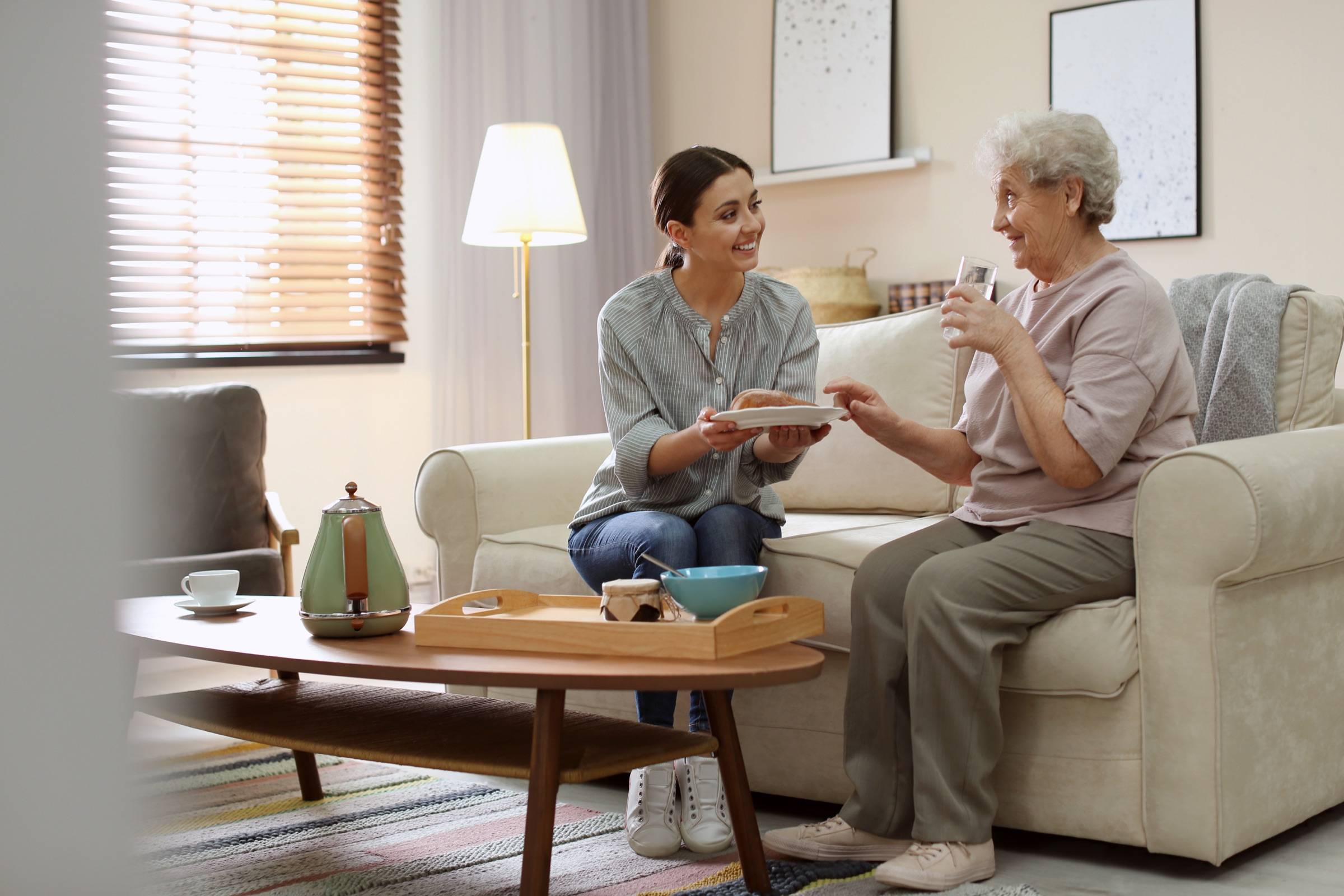 A caregiver in everyday clothes sits on the sofa with an older woman, handing her a plate as they chat and smile in her living room.